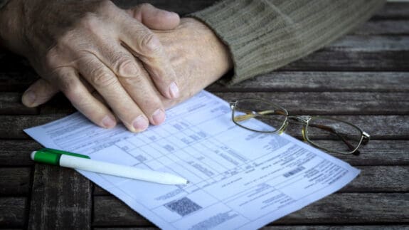 Hands of old man fills in form on wooden table.