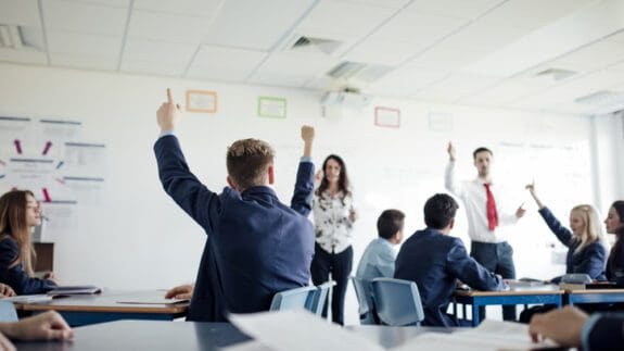 Teenagers in school uniform sitting in classroom with arms raised to answer a question