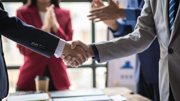 Business men shaking hands in a conference room while people applaud in the background.