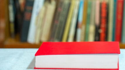 Red book on table with books on shelf in soft focus behind