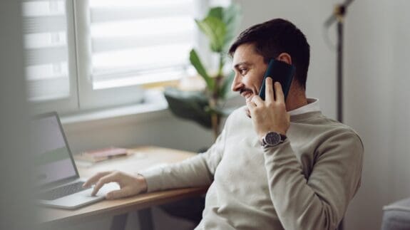 Man working on a laptop while speaking on the phone in a home office setting.