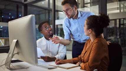 Image of 3 people working together on an audit by talking and writing notes behind a computer screen.