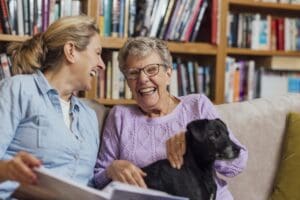 A mature woman sitting with her mother, who has dementia, and pet dog on a sofa. They are looking through a photo album together.